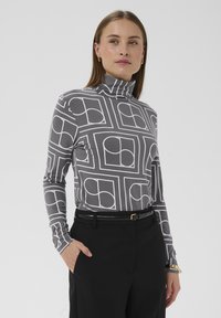 Grå langærmet rullekravebluse med hvide geometriske mønstre. Kombineret med sorte bukser og et sort bælte med en guldbæltespænde.