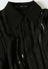 Chemise noire boutonnée avec col pointu et rayures verticales de sequins noirs sur le tissu.
