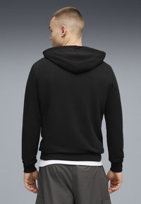 Sweat-shirt noir à capuche, en tissu doux. Présente des poignets et un ourlet côtelés, porté par-dessus une chemise blanche, vue de dos.