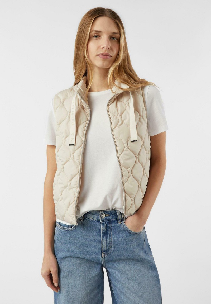 Béžová prošívaná vesta s vysokým límcem, zapínáním na zip a šňůrkami. N nošena přes bílé tričko a kombinována s modrými džínami.