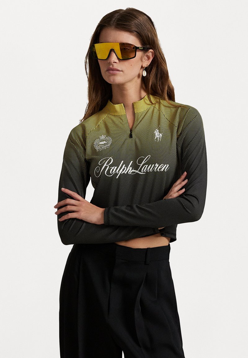 Svart og gul langermet sykkeltrøye med halv glidelås, mesh-struktur, med "Ralph Lauren"-logo og sirkulært emblem. Modell iført solbriller.