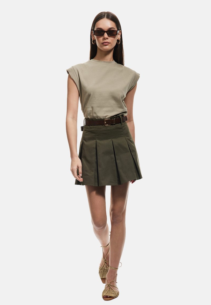 Gonna plissettata verde oliva con una cintura marrone, abbinata a un top fitted verde chiaro. La modella indossa occhiali da sole scuri e sandali con cinturini beige.