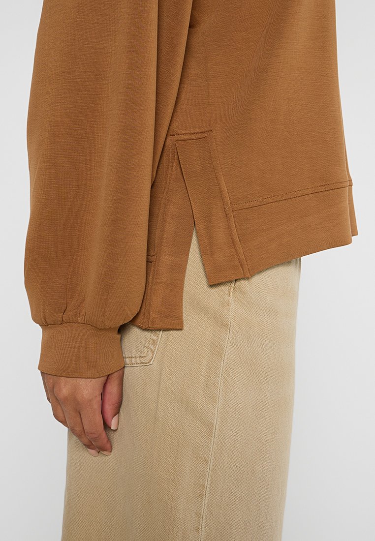 Lange bruine overhemd met getextureerde stof, zijsplitten en aansluitende manchetten, gecombineerd met beige broek. Het materiaal lijkt zacht en lichtgewicht.