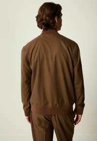 Braune Zip-Jacke aus gewebtem Stoff, mit einem gerippten Kragen und Bund. Das Design ist minimalistisch mit einer glatten Textur.