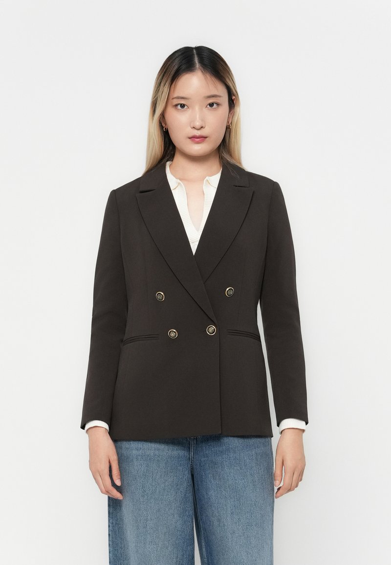 Brun dobbeltspent blazer med gullknapper, slå-krage og detaljerte frontlommer, sammen med en lyseblå bluse og blå brede jeans.