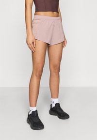 Αθλητικό σορτς σε ανοιχτό ροζ χρώμα με ελαστική μέση, σχεδιασμένο με καμπυλωτό τελείωμα και κατασκευασμένο απ ό ελαφρύ ύφασμα. Φοριέται με μαύρα αθλητικά παπούτσια.