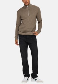 Beżowy sweter z ribbowanym wzorem, zapinany na zamek, czarne dżinsy oraz białe sneakersy z zielonymi akcentami. Krój jest luźny, a design minimalistyczny.