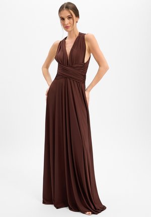 Woman wearing a sleeveless dark brown floor-length gown with a deep V-neck and ruched waistband, standing with hands on hips on white background.