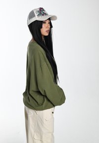 Olive zielony oversize'owy sweter z grafiką, w połączeniu z beżowymi spodniami cargo z kieszeniami. Biała siatkowa czapka z kolorowym wzorem.