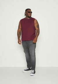 Bordeaux-nakkeless skjorte, grå jeans og svarte Converse-sneakers. Modellen har flettet hår og solbriller, stående mot en lys bakgrunn.