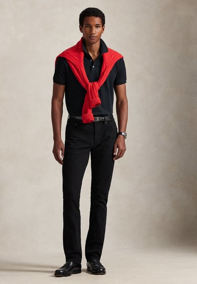 Άνδρας που φοράει μαύρο πόλο και παντελόνι με κόκκινο πουλόβερ περασμένο στους ώμους, στέκεται μπροστά σε ουδέτερο μπεζ φόντο.