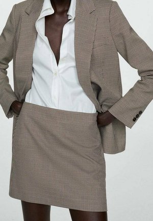 Personne portant un blazer et une jupe à carreaux marron et blanc avec une chemise blanche boutonnée, les mains dans les poches du blazer.