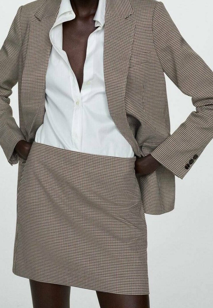 Personne portant un blazer et une jupe à carreaux marron et blanc avec une chemise blanche boutonnée, les mains dans les poches du blazer.