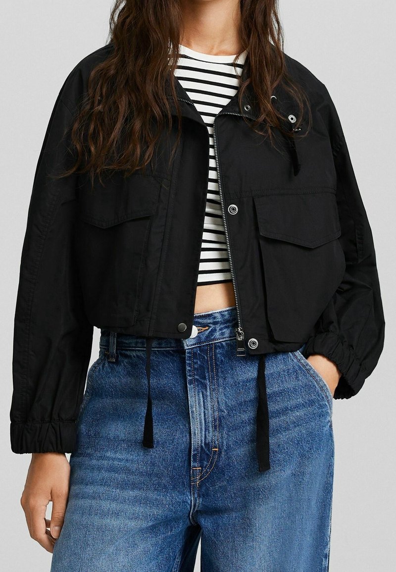 Giacca corta nera con chiusura a zip, due tasche sul petto, polsini elasticizzati e coulisse sull'orlo; indossata sopra una maglia a righe bianche e nere.