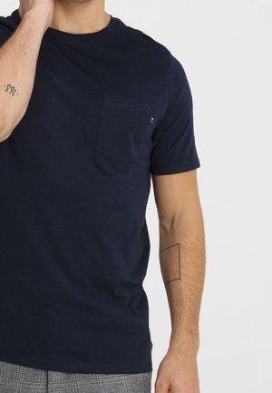 Mężczyzna w granatowym T-shircie z krótkim rękawem i kieszenią na piersi, pokazujący tatuaże na przedramionach na białym tle.