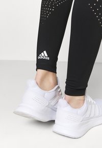 Leggings de sport noirs avec des détails perforés et un logo Adidas, associées à des chaussures de course en mesh blanches avec des languettes rembourrées et des semelles texturées.
