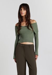 Junge Frau in einem grünen, schulterfreien, gerippten Strickpullover und hochgeschnittenen, dunkelbraunen Hosen, die vor einem schlichten Hintergrund steht.
