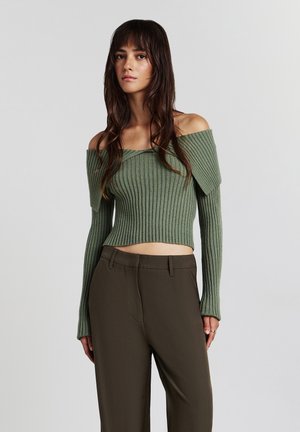 Jeune femme portant un pull en tricot côtelé vert à épaules nues et un pantalon taille haute marron foncé, se tenant devant un fond uni.