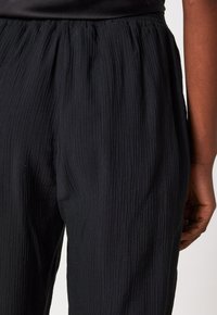 Close-up van een persoon die losse zwarte textuurbroeken met een elastische tailleband draagt, waarbij een deel van hun hand en arm zichtbaar is.