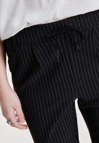 Zwarte gestreepte broek met een trekkoord in de taille, met een gladde textuur en een getailleerde pasvorm, gedeeltelijk zichtbaar onder een lichte top.