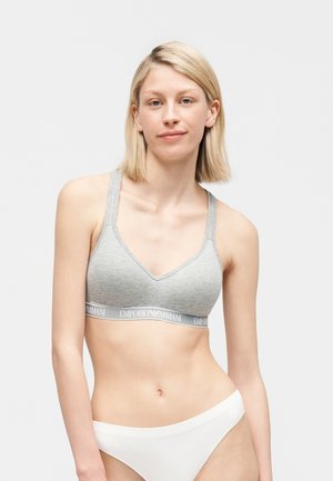 Blonde vrouw die een grijze Emporio Armani sportbeha en witte onderbroek draagt, staand tegen een effen lichte achtergrond en recht vooruit kijkend.