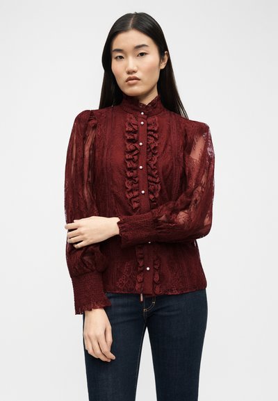 Γυναίκα με μακριά σκούρα μαλλιά που φοράει μια διαφανή μπορντό δαντελένια μπλούζα με βολάν και κουμπιά σαν πέρλες, συνδυασμένη με σκούρο τζιν.