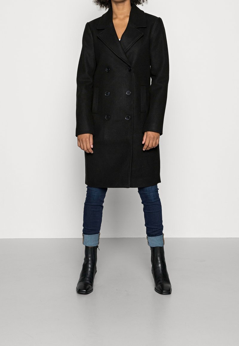 Personne portant un manteau noir croisé arrivant aux genoux, un jean bleu foncé retroussé aux chevilles et des bottines noires, debout devant un fond uni.