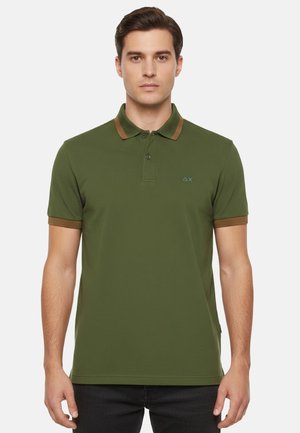 Uomo che indossa una polo verde oliva con colletto e polsini marroni, in piedi davanti a uno sfondo bianco uniforme.