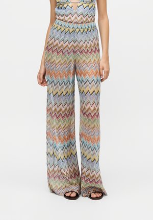 Pantaloni a gamba larga con motivo a zigzag colorato in tonalità pastello, indossati con sandali neri aperti sulle dita, su sfondo chiaro.