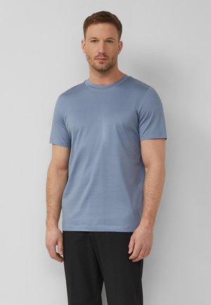 Homme aux cheveux courts portant une chemise bleu clair à manches courtes et un pantalon noir, debout devant un fond gris uni.
