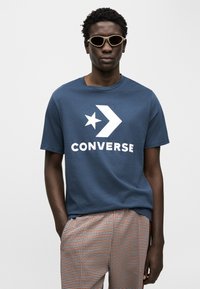 Μπλε θαλάσσης βαμβακερό μπλουζάκι με λευκό λογότυπο "CONVERSE" και σχέδιο αστεριού. Το μοντέλο φοράει καφέ παντελόνι με σχέδια και γυαλιά ηλίου ανοιχτού χρώματος.