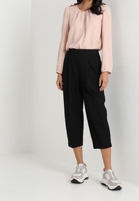 Kvinde iført en lyserød langærmet bluse, sorte cropped bukser med vide ben og hvide og grå sneakers står op ad en hvid væg.