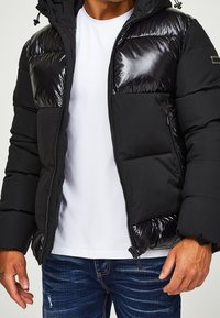 Svart puffjacka med glänsande sektioner, dragkedja, huva och sidofickor. Bärs över en vit t-shirt och i kombination med blå jeans.