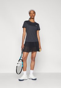 Μαύρη ριγέ αθλητική μπλούζα σε συνδυασμό με πλισέ μαύρη φούστα. Κρατούν μια ρακέτα τένις και μια μπάλα, με άσπρα παπούτσια και ελαφρύ καπέλο.