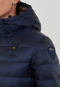 Primo piano di una persona che indossa un giubbotto imbottito blu navy con cappuccio, con etichette "Blauer U.S.A." sul petto e sulla spalla.