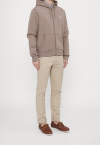 Felpa con zip marrone in tessuto morbido, abbinata a pantaloni chinos beige e scarpe di pelle marrone, che mostrano un look casual e un fit confortevole.