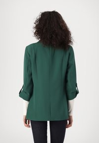 Zielony żakiet wykonany z strukturalnej tkaniny. Posiada podwinięte rękawy z akcentami guzików oraz dopasowany krój, pokazany z tyłu z gładkim wykończeniem.