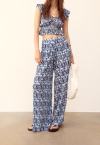 Vrouw in een blauw-wit gestreepte crop top en wijde broek, met een witte geperforeerde tas in de hand en sandalen aan.