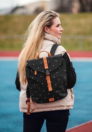 Černý látkový batoh s bílými tečkami, hnědými koženými prvky, klopovým uzávěrem a nastavovacími popruhy. Nesený na zádech osoby.