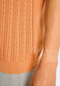 Suéter naranja de punto con patrones de trenzado y textura de tejido abierto. Presenta un dobladillo acanalado y una pequeña etiqueta adjunta en el lateral.