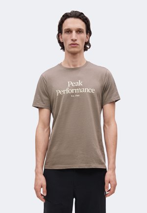 Bărbat stând în picioare, cu fața înainte, poartă un tricou taupe cu inscripția „Peak Performance Est. 1986” pe piept și pantaloni negri.