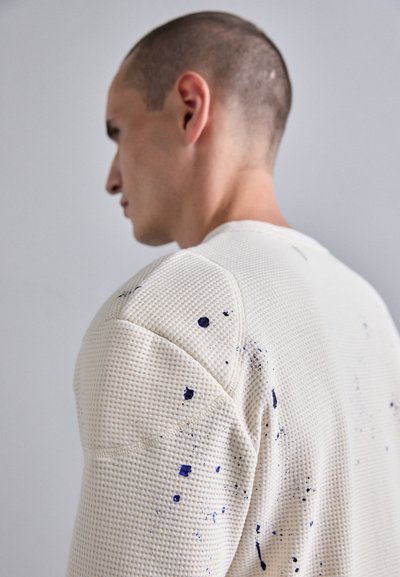 Maglione bianco texture con motivo a nido d'ape e dettagli di macchie di vernice blu navy. Scollatura rotonda e cuciture sulle spalle distintive.