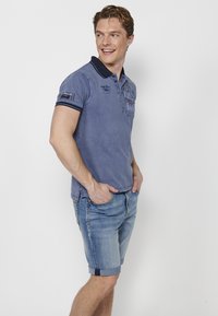 Modra polo majica z mornarsko modrimi detajli, izvezenimi grafičnimi elementi in gumbom. Upoštevane so lahke modre denim kratke hlače s zavihanimi robovi.