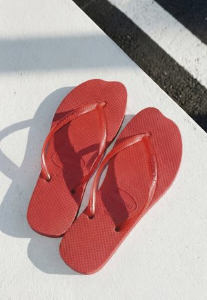 Coppia di infradito rosse poggiate su una superficie bianca con ombre, vicino a un bordo del marciapiede a strisce nero e bianco.