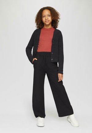 Zwarte geribbelde cardigan over een bordeaux top, gecombineerd met zwarte wijde broeken en witte sneakers. Krullend haar en handen in de zakken.