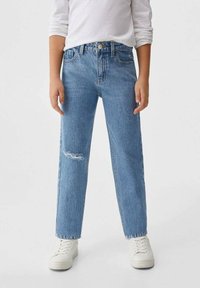 Person som bär ljusblå högmidjade jeans med en liten reva på vänstra låret, i kombination med vita sneakers och en vit långärmad tröja.