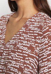 Brązowa tkanina w wzory z białymi, abstrakcyjnymi kształtami. Projekt z dekoltem w serek, miękka faktura i luźny krój, koncentrujący si�ę na górnej części klatki piersiowej.