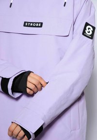 Lavendel vandtæt jakke med en frontlomme, prydet med et sort logo-patch på ærmet og justerbare håndledskanter med Velcro-lukning.