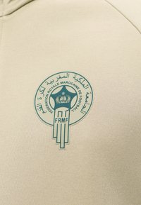 Έμβλημα της Fédération Royale Marocaine de Football με ένα στέμμα μέσα σε ένα αστέρι και αραβικό και γαλλικό κείμενο σε μπεζ ύφασμα.