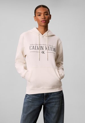 Frau trägt einen hellbeigen Calvin Klein Hoodie mit vorderen Taschen und blauen Jeans vor einem schlichten grauen Hintergrund.